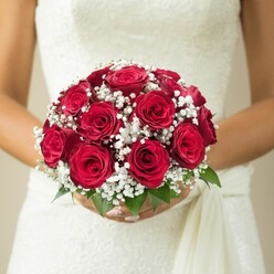 Nestled in the heart of Canonbury near the quiet, leafy paths of Compton Road, a bride's bouquet rests in gentle hands, radiating an air of traditional romance. Tightly clustered, deep red roses form the rich, lustrous core of this arrangement, each bloom unfurling with velvet softness and subtle perfume. Snowy clusters of baby's breath, their blossoms minute and airy, float delicately among the roses, bringing a touch of ethereal beauty and creating striking contrast against crimson petals. Bright green leaves peep from the base, grounding the bouquet with a hint of vibrant freshness that brings to mind Canonbury Gardens on a spring morning. The bride's manicured hands cup the bouquet lovingly at her midsection, her white wedding gown's textured lace bodice and flowing sash softly visible behind, evoking the artisan charm of local floristry. Soft, natural light washes the scene, ensuring every petal and fabric thread is alive with subdued radiance. This image celebrates not only the enduring allure of the classic rose bouquet but also the distinctive artistry of Canonbury's local florists-making it an ideal visual for wedding planning stories, reflections on bridal style, or tales of romance blossoming in North London.