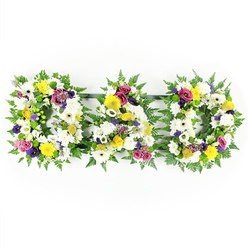 A beautifully composed funeral tribute spells out DAD in three bold floral letters, laid out in a neat row on a spotless white background so every colour reads clearly. Each individual letter is framed with a thick collar of glossy green fern, the feathery leaves giving a soft, garden-like feel and providing a strong outline for the shapes of the D, A, and D. Inside this leafy border, the letters are tightly filled with white chrysanthemums and wide-faced white gerbera daisies, creating a cushion of velvety white petals that suggests peace, purity, and quiet farewell. Nestled among the whites, clusters of yellow gerberas and petite yellow spray roses light up the tribute with a hopeful brightness, like the warmth of shared memories. In between, accents of purple lisianthus, deep pink or fuchsia spray roses, and small lavender-toned flowers weave through the design, offering subtle drama and conveying enduring love and respect. Sprigs of bright green button chrysanthemums and other soft green touches sit between the blooms, adding freshness and depth so the letters feel full and three-dimensional. Slim, dark support rails link the three designs, but they recede into the background, keeping the visual focus on the flowers and the heartfelt word they form. This arrangement feels perfectly suited to a Canonbury farewell, perhaps resting beside family tributes after a service near Highbury Fields or along a quiet church aisle, expressing gratitude and remembrance for a cherished dad in a way that words alone often cannot.
