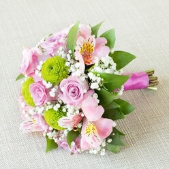 Evoking the tranquil grace of Canonbury's tree-lined streets, this exquisitely arranged bouquet rests serenely upon a light, subtly patterned linen, as if awaiting its moment in a refined North London townhouse. The composition is a harmonious medley of soft pink roses, their petals infused with gentle romance and the sweet scent of dawn, mingling with the youthful energy of lime green button chrysanthemums. Each chrysanthemum forms a cheerful pom-pom, their spirited green recalling the lush lawns near Compton Terrace Gardens. Interwoven are delicate pink alstroemeria, their inner petals painted with whispers of gold and flecks of deep red, imbuing the arrangement with visual intrigue and gentle movement. Interspersed throughout, airy clouds of white gypsophila recall dew-laced mornings along Canonbury Road, lightening the bouquet and drawing every blossom into a careful balance of colour and form. Lush green foliage, shaped by the steady hands of a Canonbury florist, anchors the arrangement with verdant depth and authentic freshness. The bouquet's stems are gathered with thoughtful precision and tied with a satin ribbon the shade of late spring azaleas, its magenta glow bringing a vibrant finish. Every detail-down to the glints of soft sunlight and the tactile joy of petal and leaf-conveys a sense of celebration, perfect for weddings, anniversaries, or a thoughtful gift delivered by Canonbury Flowers. This scene whispers of cherished memories, garden parties, and everlasting affection beneath the canopy of North London's most charming neighbourhood.