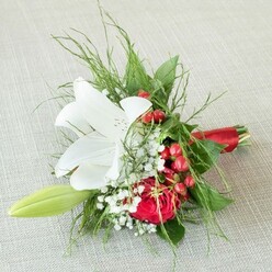 Captured from above upon a pale, woven fabric reminiscent of the classic linens found in Canonbury drawing rooms, this artful bouquet radiates understated luxury. The eye is instantly drawn to the fully bloomed white lily, its petals wide and pure, feathered with subtle chestnut speckles and nested yellow stamens that impart a warm, honeyed glow. Nestled nearby is a red rose, plush and richly layered, swirling inwards in a spiral akin to the winding walkways off Upper Street. Between these signature blooms, baby's breath whispers in lacy clusters, softening the composition with a cloudlike grace, while bright red berries glisten-harbingers of festive cheer and reminders of local winter markets. The gentle arc of a pale green lily bud hints at growth and fresh starts, echoing the community's creative spirit. Fern-like greenery and sturdy, dark leaves provide lush support, framing the flowers and highlighting their colours against the calm light. The bouquet's stems are gathered with a gleaming red satin ribbon, a touch of sophistication and warmth familiar to those who cherish special occasions here in Canonbury. This arrangement, perfect for bridal parties or meaningful gifts, radiates a feeling of intimacy and celebration, fusing the best of local floral artistry with a sense of timeless romance found in Islington's heart.