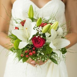 This captivating bridal bouquet, carefully embraced by a bride dressed in a stunning white gown with subtle textured patterns, tells a story of romance woven with every stem. Canonbury's floral traditions are honoured here, with large, snowy oriental lilies unfurling alongside tight, passionate red roses-each flower carrying the energy of a marriage celebrated on a peaceful Canonbury Crescent morning. Unopened green lily buds peek out, their fresh anticipation mirroring the excitement of a wedding day, while billowy sprays of tiny white baby's breath lend a dreamy, almost whimsical texture. Flashy red hypericum berries and playful button-like green blooms add festive notes, reminiscent of late-summer walks through the quiet back gardens of Canonbury Square. The entire bouquet feels balanced atop a weave of glossy and feathery greenery sourced from local growers, giving it structure and life. Set against the bride's elegant hands and a softly diffused background, every element-colourful and textural-draws the eye and uplifts the heart, speaking of devotion, joy, and the artistry found at Canonbury Flowers. This bouquet is a perfect choice for couples seeking wedding flowers that not only dazzle the eye but capture the spirit of a special day spent in one of North London's most cherished neighbourhoods.