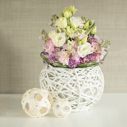 On a luminous white tabletop in the heart of Canonbury, an artistically composed spring bouquet radiates gentle sophistication. The main blooms-soft white and buttery yellow eustoma, their petals exquisitely layered-sit alongside vertical accents of pale pink matthiola, lending a sense of movement and refined height that echo the graceful facades along Canonbury Square. Dotted amidst the primary flowers, clusters of lively purple filler-reminiscent of allium or aster-impart texture and vibrancy, their hues deepening the bouquet's pastel palette. A sturdy fringe of deep green foliage frames the arrangement, grounding the lightness above. The flowers are elegantly housed in a spherical white vase, its open-work crafted from fine rattan or delicately painted branches, drawing the eye with its modern, lace-like softness and artisanal craftsmanship. To the side, two matching decorative orbs, fashioned from light, natural rattan, extend the theme of intricate weaving, suggesting homemade charm and creative spirit so beloved in Canonbury's artistic circles. The warm, subtly textured greige wall in the background diffuses the scene in tranquil, filtered light, allowing the bouquet's colors and textures to glow. Evoking the renewal of spring and the communal warmth of North London gatherings, this arrangement is the essence of Canonbury floristry, perfect for weddings, celebrations, or as an elegant statement in a stylish home.