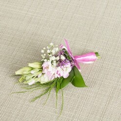 A single, elegant boutonniere or small corsage is laid carefully on a pale beige, woven linen surface, the fabric's fine texture giving the scene a calm, understated backdrop reminiscent of a relaxed Canonbury townhouse reception. The floral arrangement is composed around a tight cluster of soft pink, multi-petalled blooms, reminiscent of miniature stocks or tiny garden roses, their petals gently ruffled and overlapping like folded silk, with faint blush tones deepening towards the centres. Interspersed among these pastel flowers are delicate sprigs of crisp white baby's breath, each tiny bloom adding a cloud-like softness and an airy, romantic feel. Here and there, small spikes of lavender or deep purple florets peek through, providing a graceful contrast to the pink and white palette, much like the subtle pops of colour you might notice strolling past local gardens off Canonbury Square in spring. To one side, several slender, creamy white to pale green buds, likely freesia or a similar bloom, stretch gracefully outward, still closed, suggesting the quiet anticipation of a ceremony yet to begin. The flowers are nestled into a base of broad, glossy green leaves, rich and vibrant, with fine strands of feathery greenery and grass-like fronds drifting out from beneath, giving a gentle sense of movement. The stems at the bottom are neatly gathered and tightly wrapped with a striking fuchsia satin ribbon, finished with a precise bow whose loops and tails lie smoothly against the fabric. Soft, even lighting enhances every texture-from the satiny petals to the ribbon sheen-creating an impression of fresh, handcrafted detail, perfect for a groom's lapel at a Canonbury wedding, a prom corsage collected near Essex Road, or any intimate celebration where subtle floral artistry matters.