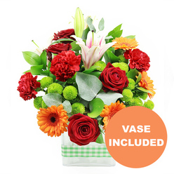 In this detailed product image, a lush, densely packed bouquet seems to burst from a sleek rectangular glass vase, every bloom carefully positioned to draw the eye. At the centre, several deep red roses, with tightly furled, velvety petals, offer a classic focal point, their rich colour echoing the brick tones of Canonbury's terraces. Interwoven with them are red-orange carnations, the ruffled edges of their petals creating a sense of movement and layered texture, as though the bouquet is in gentle motion. Radiant orange gerbera daisies with dark brown or almost black centres sit slightly forward, their flat, symmetrical faces bringing a joyful, contemporary note that would brighten any table off Upper Street. Towering above, white Oriental lilies rise on strong stems; some are fully open, revealing pink-tinged throats and prominent stamens, while others remain in tight buds, adding a vertical rhythm and promise of continued blooming. Between the larger flowers, small, round lime-green pompom chrysanthemums nestle like bright beads, their compact, cushion-like forms offering a fresh, zesty contrast. Rich green foliage, including broad, glossy leaves and sprigs of silvery eucalyptus, surrounds and supports the blooms, creating layers of depth and a natural, garden-fresh feel. The stems are clearly visible through the transparent sides of the modern rectangular vase, which is wrapped near its base with a crisp light green and white gingham ribbon, giving a hint of country charm amid the city setting. In the bottom right corner of the image, a bold circular orange overlay displays the words VASE INCLUDED in clean white capitals, clearly indicating that this chic container is included, making the arrangement an effortless, ready-to-enjoy gift for birthdays, congratulations, or welcoming a new neighbour in Canonbury.