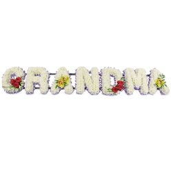 Displayed against a clear, uncluttered white background, this image shows a lovingly constructed funeral flower tribute spelling the word GRANDMA in large block capitals, the kind of design often chosen by families across Canonbury to rest beside the coffin at a final farewell. Each letter is thickly padded with tiny, perfectly opened white chrysanthemum heads, pressed so close together that no gaps of greenery show, creating a plush, velvety texture like a soft quilt of petals. Along the lower edge of every letter runs a slim border of pale lavender flowers, perhaps statice or small asters, their cool purple tones lending gentle contrast and outlining the shapes with a refined, almost lacy finish. The tribute is further personalised with carefully placed accent blooms. On the lower left curve of the G, three rich, deep red roses sit among dark green leaves, their colour symbolising heartfelt devotion and respect. The first A is decorated with two yellow-green orchids set along the crossbar, their unusual colour and neat, waxy petals adding an elegant touch of individuality. The R, N, and D remain pure white within their lavender frames, allowing the eye to rest before reaching the M, where another cluster of three red roses nestles into the lower right section. The final A echoes the first with another pairing of the same distinctive green orchids. A dark green or black metal stand runs behind the letters, holding them in a straight line and making the tribute suitable for display at a chapel, crematorium, or graveside near Canonbury's quiet residential streets. Overall, the arrangement conveys peace, tenderness, and family pride, a soft yet striking memorial to a cherished grandmother.