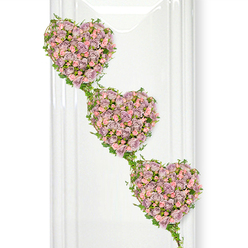 Three interlinked hearts of roses cascade softly down a clean white background, forming an elegant funeral tribute that feels both contemporary and timeless. Each heart is shaped entirely from clusters of miniature and small rose blooms, predominantly in a muted lavender or dusty mauve shade, blended with lighter pastel pink roses that introduce warmth and tenderness. The flowers are placed closely together, so that the hearts appear plush and almost cushion-like, with layers of velvety petals creating depth and subtle shadow. The arrangement descends diagonally from the top left, giving the sense of a gentle, flowing journey, perhaps mirroring the passage of a loved one's life. Fine strands of fresh ivy weave around and between the hearts, their vibrant green leaves looping delicately over the curves, symbolising connection and unbroken bonds. The ivy acts as a natural frame and stem, guiding the eye from one heart to the next and uniting them as a single composition. The background is bright and uncluttered, with faint vertical reflections that suggest glass or a polished wall, allowing the soft purple and pink tones to stand out with clarity and calm. This thoughtful design would be at home in a Canonbury memorial service, offering a quiet statement of love and gratitude that feels deeply sincere. While there is no visible vase or stand, the structured nature of the hearts reveals skilled floral craftsmanship, reflecting the kind of careful, local attention families in Canonbury often seek when commemorating someone special.
