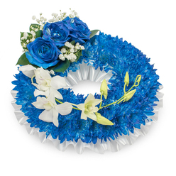A dignified circular memorial wreath photographed on a plain white backdrop, its body densely covered in vivid, deep blue chrysanthemum-like flowers that form a sumptuous, continuous ring. The tiny petals of the chrysanthemums create a richly textured surface in sapphire and royal blue tones, while a pleated white satin ribbon subtly lines the inner opening and outer edge, lending a polished, ceremonial border. At the top-left curve a trio of large blue roses unfolds in full bloom; their layered, velvety petals register a slightly darker azure hue that harmonises with the chrysanthemum base. Around these roses, soft sprays of baby's breath and a few dark-green leaves add a gentle, classic contrast and an understated green depth.

A sweep of pure white orchids-suggestive of Phalaenopsis petals-cascades from the mid-left across the wreath toward the lower-right, their smooth, paper-like petals creating an elegant, whispering movement. Sprigs of fresh green orchid buds punctuate the cascade, some tight and others beginning to open, speaking quietly of continuity and new growth amid remembrance. The palette of deep blue and pristine white, combined with mixed textures from velvety to silky, gives a calm, reverent impression ideal for funerals or memorials near Canonbury and along New North Road. Prepared with local care and attention to floral balance, this wreath can be arranged by Canonbury Flowers for services in the neighbourhood or nearby chapels.