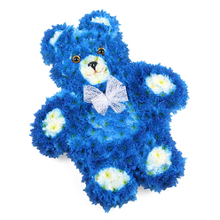 Set against a clean white background, this floral teddy bear tribute appears almost to float, an intricate sculpture woven entirely from fresh blooms in intense royal blue and soft, creamy tones. The body is densely covered in small blue flowers, each head tucked close to its neighbour so the surface looks like a luxurious, plush fabric, with gentle ripples around the rounded belly, curved arms, and slightly splayed legs. Along the inner ears, the rounded muzzle, and the four oval paw pads, a cluster of pale cream or off-white blossoms provides tender contrast, like patches on a favourite toy that has been loved for years. The face is expressive yet peaceful: deep amber-brown eyes with dark pupils shine softly, set above a neat black triangular nose and a fine curved line that hints at a gentle, reassuring smile. Around the neck, a delicate white organza-style bow is tied with graceful loops and trailing ends, its translucent fabric catching the light and softening the strong blue tones. The bear is positioned on a slight diagonal, giving a feeling of movement, as if it has just been laid down with care. As a florist working in and around Canonbury, where families often seek something uniquely tender for services in local chapels and homes, I see this design as a deeply personal funeral tribute or memorial for a child, or a baby boy's remembrance. The royal blue flowers suggest calm, loyalty, and eternal love, while the creamy highlights and hand-crafted details bring warmth and intimacy, making this floral teddy ideal for heartfelt deliveries throughout Canonbury and neighbouring parts of North London.