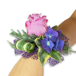 A gentle halo of floral textures and evening-tinged colour defines this wrist corsage, where classic romance meets subtle modernity. The focal rose opens in a tender mauve-pink, its layered petals catching a faint, satiny light as if still holding morning moisture; its tone carries a soft blush that deepens toward the outer ruffle. Nestled close are rich, saturated hydrangea clusters in indigo-blue and deep violet, their tiny florets offering delicate points of light at each centre and creating a calm, twilight mood. Variegated English ivy, with dark green hearts and creamy margins, threads through the arrangement, offering contrast and a cool, glossy texture. Behind the flowers, slim silvery foliage-suggestive of eucalyptus or lavender-fans out in feathery strokes, adding breath and a soothing, aromatic lift. A single broad leaf striped in white and green is artistically curled into a narrow spiral, providing a neat, contemporary flourish that keeps the composition feeling fresh and tailored. Beneath all, an airy deep-purple mesh cradles the blooms, introducing a playful touch of lace-like texture and subtle depth. The corsage sits on a light-toned forearm against a bright white field, which makes the purple hues sing. As a local florist mindful of occasions in Canonbury and nearby Newington Green, I see this as perfectly suited to bridesmaids or honoured guests-an accessory that smells faintly of green summer and is crafted to be remembered after the last dance.