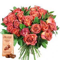 With the exquisite craftsmanship of a Canonbury florist, this stunning rose bouquet embodies both artistic flair and deep local charm. Rows of open roses bloom in generous abundance, their petals swirling outward in rich gradients: sunset coral, blush peach, warm salmon, and sultry pink merging together, like the sky over neighbouring Islington at dusk. Each flower varies subtly in tone, some revealing creamy yellow or gentle ivory at their centres, gradually flushing into more saturated pinks and reds. The lush green foliage - perhaps freshly gathered eucalyptus and glistening salal - envelops the blossoms, adding both vibrancy and a crisp, woodland fragrance reminiscent of walks under the trees of Compton Terrace Gardens. Alongside the bouquet, a premium Thorntons Orange Crisp chocolate pouch in a pale beige hue stands upright, its golden lettering highlighted by the light. The packet is adorned with an inviting illustration of two chocolate truffles, while just ahead, real truffles rest, their dark, glossy exteriors hinting pleasantly at orange and silky caramel. Every detail in this composition is curated to evoke the feeling of celebration - a gift that brings comfort, romance, and a taste of luxury to occasions like Mother's Day, anniversaries, or birthdays. Delivered fresh and swiftly throughout Canonbury, this floral and chocolate duo creates an experience to be savoured with every sense.
