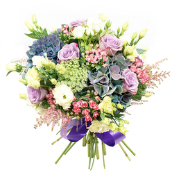 The essence of Canonbury's tranquil charm is captured in this meticulously arranged bouquet, where every bloom speaks to artful design and local character. At its core, plush hydrangeas in rippling waves of blue, teal, and velvety violet set a dramatic, calming tone, their fullness reminiscent of the lush gardens tucked behind Canonbury's historic terraces. Among them, roses in gentle lilac and creamy white unfurl gracefully, their petals imbued with the quiet romance of these north London streets. White ranunculus, dense and velvety, and the pale yellow buds of eustoma punctuate the arrangement, their shapes lending lightness-as if touched by morning sun streaming through the windows of Georgian homes. Sprigs of vivid pink filler flowers, paired with feathery astilbe, wind throughout, bringing energetic accents and evoking the layered textures of New River Walk's blossoming borders. Stems of lacy filler foliage, almost translucent, add a subtle, intricate touch, while deep green leaves offer a rich backdrop that underscores the bouquet's vibrant colours. Gathering every stem together, a satin ribbon in royal purple envelopes the base, adding an elegant finish and reflecting the creative spirit of Canonbury's local florists. Designed as a centerpiece for celebrations or as an uplifting gesture delivered locally, this arrangement radiates peace, joy, and the singular artistry found only in a neighbourhood florist's touch.