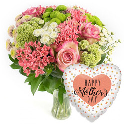 A bright, celebratory Mother's Day gift arrangement is shown in a clean studio setting, the pure white backdrop echoing the light townhouses and leafy calm of Canonbury's residential streets. At the centre of the image stands a rounded bouquet in a clear glass vase, brimming with fresh blooms that lean toward soft springtime tones. Several large pink roses form the heart of the design, their petals opening in generous spirals, tinted with deeper pink toward the centre and softening to delicate, almost cloud-like edges. Around them, clusters of coral-pink blossoms, similar to kalanchoe, create a lively, textural ring, each tiny flower catching the eye with its compact, starry shape. Lime green button chrysanthemums nestle among the pinks, their spherical, almost tufted heads adding a modern, slightly quirky contrast that feels both fun and elegant. Sprays of small white, daisy-like chrysanthemums weave through the composition, bringing lightness and balance, while rich green foliage fills out the base, giving a sense of natural abundance. The stems, visible through the vase, are neatly cut and immersed in clear water, reinforcing the impression of freshness you'd expect from a same-day Canonbury delivery. Just to the lower right of the bouquet floats a lustrous heart-shaped foil balloon. Its white or pale silver background is dotted with irregular polka dots in gold, silver, and soft peach, like confetti drifting through the air. At its centre sits a solid peach-coral heart carrying the phrase "HAPPY Mother's DAY" in elegant black lettering. Together, the flowers and balloon convey warmth, appreciation, and happy anticipation, as if ready to be carried up a narrow Canonbury terrace staircase and placed on a kitchen table for a well-loved mum.