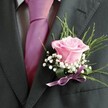 A poetic arrangement unfolds on the lapel of a sharply dressed figure ready for a celebration somewhere along the peaceful avenues of Canonbury. At its centre, a blush-pink rose opens with the quiet elegance of a heartfelt promise, its velvet-soft petals curving softly, as if to cradle a secret. Around the rose, fine sprigs of white baby's breath create a dreamy, cloud-like frame, enhancing the bloom's tender hue. Wisps of deep green foliage adjust behind, their organic lines adding movement and recalling the gentle wildness of local parks along the Regent's Canal. The suit jacket-a deep charcoal, textured and refined-anchors the florals, while a shimmering lilac-fuchsia tie provides a playful, yet sophisticated note atop a subtly patterned grey shirt. The look is pulled together by a silken lavender ribbon at the base, complementing the composition while hinting at joyous moments yet to come. For a wedding or civil union near Highbury Fields or at one of Canonbury's beloved town halls, this buttonhole becomes more than an accessory: it's a tactile, aromatic keepsake, crafted with skill and care to infuse a sense of occasion, anticipation, and belonging right at the heart of North London.