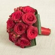 Resting gently atop a soft, neutral-toned linen-like the crisp fabrics seen in Canonbury's weekend markets-this compact bouquet of deep red roses exudes timeless romance for heartfelt weddings and cherished moments. Each of the ten showy roses is in perfect, open bloom, flaunting unfurled, velvet petals in bold crimson hues, while bright red hypericum berries nestle among the flowers, adding playful punctuation of festive colour and subtle contrast. The effect is vibrant yet elegant, the arrangement a living painting crafted in Canonbury's renowned floristry style. Encircling the blooms, lush broad leaves of profound emerald provide a sculpted green backdrop, gently folded to enhance the roses and frame the berries. The stems are neatly gathered and finished with a polished, scarlet satin ribbon that peeks out at the base, uniting the bouquet with understated sophistication. Even, diffused light lavishes the entire scene, bringing out the petal textures, glossy berries, and the smooth sheen of the ribbon without casting harsh shadows. Scented with the dusk-like perfume of fresh roses, this arrangement embodies passion and tradition, perfect for Canonbury couples saying I do in hidden gardens or gifting expressions of love that linger long after the moment passes.