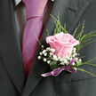 Pinned neatly to a charcoal suit, this blush-pink rose boutonniere exudes the gentle poise of a Canonbury ceremony, perhaps in a townhouse near New River Walk. The single rose is the clear focal point, its petals unfolding in a soft spiral of pale pink that deepens only slightly toward the edges, creating a calm, romantic glow. The surface of each petal looks velvety and touchable, with a subtle play of light that reveals their fine texture rather than a glossy shine. Nestled around the rose are tiny, cloudlike sprigs of gypsophila, each white bloom no larger than a pinhead yet gathered into airy clusters that add volume without weight. Fine green foliage - slender, fresh, and lightly arched - cradles the composition, suggesting a sense of breath and movement, like leaves stirred by the breeze that drifts along the quiet streets of Canonbury and into nearby Islington Green. At the base, a pale mauve or soft lilac ribbon is wrapped and tied with deliberate care, its satin finish catching the light just enough to signal thoughtful attention to detail, a small echo of the silk tie beside it. The interplay of textures is refined: the smooth suit fabric, the plush rose petals, the tiny, textured gypsophila, and the polished ribbon all working together to create a harmonious, understated elegance. The lighting is soft and flattering, casting gentle highlights along the rose's curves and lending the greens a fresh, dewy look. This boutonniere feels designed not only for style but for emotion - perfect for a first dance in an intimate Canonbury venue or a walk home under the trees after the celebration, the rose still fresh and lightly scented as the evening quietens.