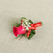 Laid diagonally against a subtle beige fabric that resembles fine canvas, this boutonniere reads like a small study in colour and texture crafted for memorable occasions. A bold magenta rose claims the centre, its petals tightly furled at the heart and softly opening outward in a velvet spiral that invites touch. Nestling around the rose are clouds of delicate Gypsophila (baby's breath), their tiny white blooms lending an airy, nostalgic softness that balances the rose's saturated tone. Scattered just behind the main bloom, bright red hypericum berries add warm highlights, their glossy skins reflecting light and echoing the ribbon's deep hue. Slim, wispy blades of fern-like greenery provide movement and a wild, natural silhouette, contrasted by larger, folded aspidistra leaves that give structure and depth to the design. Every stem is expertly wrapped in a crimson satin ribbon, tightly bound and finished with a neat knot that keeps the composition secure while adding a considered, polished flourish. The linen-like background and gentle morning light bring out the tactile differences-the rose's velvet, the berries' gloss, the grasses' whisper-while the scent suggests a soft rose perfume with fresh green notes. As a florist familiar with Canonbury and nearby New River Walk, I imagine this pinned to a lapel at a wedding on a balmy late-summer afternoon, where it would read as both romantic and subtly spirited.