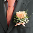 This boutonniere is a study in gentle contrast and thoughtful restraint, handcrafted with the kind of quiet care you expect walking down Upper Street toward Canonbury Square. A single perfect peach rose commands the centre, its blush-toned petals layered like a sunrise - creamy at the edges, warmed with coral-pink at the core - and catching ambient light to create a soft, hopeful glow. Surrounding the bloom are vapour-like clusters of white baby's breath (gypsophila), their tiny blossoms drifting around the rose like celebratory confetti or a halo of innocence. Slim, fresh green foliage and a curled leaf give structural grace and a suggestion of wildness, their cool, verdant scent balancing the rose's faint sweetness. Everything is bound at the stem with a satin ribbon that mirrors the tie's warm coral-pink sheen, tied into a modest bow that speaks of precision and personal care. Against the charcoal jacket, the colours resolve into an elegant, contemporary palette - sophisticated yet warmly intimate, suitable for a groom, best man, or a special guest. Texturally the piece is invitingly tactile: the velvety rose, the airy gypsophila, the crisp ribbon and feather-light fern. Created with flowers sourced fresh from our Canonbury workshop and finished with neighbourhood sensibility, it feels at home in quiet church halls, riverside ceremonies near Essex Road, or an intimate reception at a local tavern. The overall impression is one of promise and reassurance, a small floral statement that whispers love and celebration rather than shouting it.