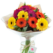 A vividly coloured bouquet of hand-tied Gerbera daisies is captured in radiant studio light, standing proud in a clear glass vase, with the fresh, verdant stems and water glinting through. The daisies' petals flare outward in shades of golden yellow, effervescent orange, and lush, deep fuchsia-an exuberant palette that brings to mind the vibrant community gardens of Canonbury and the lively stalls of Newington Green market. Each bloom features a pronounced, nearly black centre, creating a striking focal point amidst the swirl of colour. Delicate stems of greenery are nestled within, providing a natural texture and balance to the flowing forms. The bouquet is lovingly wrapped in rustic brown kraft paper, a texture that echoes the charm of local delis, and finished in a clear, glossy cellophane layer shaped into a classic protective cone. A band of natural twine ties the stems securely within the vase, from which a small, white, rectangular tag-embellished with a red rose illustration-hangs, suggesting a personal message or special occasion. This bouquet radiates cheerful abundance and refined simplicity, ideal for anyone in Canonbury looking to share a gesture of kindness, celebrate a milestone, or display a handcrafted piece of floral artistry within their home.