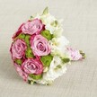 A lush, rounded bridal-style bouquet is photographed from a slightly raised angle, resting on a softly textured light beige fabric that gently diffuses the light, much like an overcast morning in Canonbury. At the centre, large pink roses in full bloom steal the eye, their petals swirling from a tender lavender blush at the heart to a deeper, almost fuchsia-pink on the outer edges, giving a romantic two-tone effect that feels rich yet refined. Threaded between these statement roses are clusters of small chartreuse green button chrysanthemums, their tight, domed heads adding playful texture and a crisp, modern pop of colour that keeps the design feeling fresh and contemporary. Delicate white flowers, likely lisianthus, cradle the brighter tones, their soft, layered petals and pale greenish-yellow centres bringing a cloud-like lightness, as if catching the last glow of daylight over nearby New River Walk. A few closed buds are tucked in, suggesting the bouquet was arranged at peak freshness, ready for delivery across Canonbury. The stems are neatly gathered and wrapped in smooth pink satin ribbon, forming a graceful handle that would rest comfortably in the hand. Over this, a slender strand of tiny white pearls spirals down, echoing vintage bridal jewellery and adding a subtle sheen. The overall mood is one of romance and celebration, ideal for an intimate Canonbury town hall wedding, an elegant engagement dinner on a local terrace, or a luxurious gift that conveys heartfelt emotion with understated sophistication.
