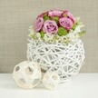 A stylish floral centerpiece rests on a spotless white tabletop, capturing the calm, contemporary charm often found in Canonbury homes. At its heart is a round, openwork white vase, woven from rattan-like twigs that intertwine into a light, spherical form, allowing glimpses of the stems within and creating a subtle lattice of soft shadows. From this sculptural base rises a dome of pastel flowers: velvety roses in shades of gentle pink and whispering lavender, some tipped in richer pink along the outer petals, open fully to reveal intricate spirals of colour. Between them, bright green button chrysanthemums add crisp dots of texture, their neat, globe-shaped blooms bringing a fresh, almost playful contrast to the romantic roses. Around the lower edge of the arrangement, tiny white filler flowers cluster and drift slightly over the rim, giving the bouquet a relaxed, natural finish rather than a rigid outline. To the left, two decorative spheres made from pale wicker or light wood sit side by side on the same white surface, one larger and one smaller, their open, woven patterns echoing the vase and reinforcing the crafted, organic feel. The softly blurred background in warm beige tones, reminiscent of a linen curtain or textured wall, keeps attention squarely on the flowers while adding gentle warmth. The overall impression is of a refined wedding table arrangement, the kind a local Canonbury florist might create for a reception near New River Walk or a small celebration tucked away on a quiet residential street, radiating softness, romance, and thoughtful design.