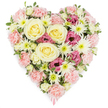This image shows a richly textured, heart-shaped floral tribute set crisply against a pure white background, designed to convey compassion, remembrance, and enduring love. The entire heart is packed with blooms, creating a lush, unbroken surface of colour and petal. At its core, several large roses in creamy yellow to soft ivory gently open, their tightly swirled petals suggesting warmth, gratitude, and the lasting bond between the departed and those who remain. Surrounding these focal roses, pastel pink carnations form a soft halo, their frilled, voluminous petals overlapping in dense layers that trace the curve of the heart and lend a comforting, padded effect. Dotting the arrangement are white spider chrysanthemums, their fine, radiating petals and fresh green centres adding airy texture and a sense of light and clarity within the design. Tucked among the paler shades, deeper magenta-pink lisianthus blooms provide a richer accent, their gently ruffled, bell-shaped flowers creating pockets of depth and visual interest. Small, variegated green leaves peek out in subtle flashes of foliage, reminding the viewer of the freshness and living quality of the tribute. At the pointed tip of the heart, a slender pink ribbon is just visible, indicating this piece has been crafted as a sympathy or funeral arrangement, perhaps for a farewell in Canonbury, a service near Canonbury Station, or a memorial gathering by the leafy streets around Alwyne Villas. The overall feeling is of a carefully considered floral heart, created to offer comfort, honour cherished memories, and speak softly where words feel insufficient.