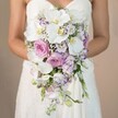 Set against a gently blurred, warm beige background reminiscent of Canonbury's tranquil architecture, this close-up photograph reveals a bride from the mid-chest downward, her arms gracefully supporting a luxurious cascading bouquet. The arrangement pours down in an elegant teardrop shape, its dominant white phalaenopsis orchids instantly drawing the eye, their wide, silky petals and sunny yellow centers exuding quiet luxury. Around these, lush blush and light mauve garden roses billow with softness and romance, invoking the scent and feel of blossoming parks in Canonbury during spring. Interspersed are delicate lavender-toned stock and lisianthus-some flowers edged in soft purple for visual intrigue-complemented by scattered sprigs of airy white gypsophila and subtle hints of greenery. Every flower is hand-placed to create a sense of airy abundance and gentle movement, reflecting expert craftsmanship and a deep understanding of balance. The bride's strapless white gown, laced with fine embroidery at the bodice, peeks elegantly beneath the bouquet, its texture harmonizing with the intricate blooms. Together, this scene radiates timeless romance and sophisticated celebration-ideal for weddings in the heart of Islington or beside Canonbury's peaceful gardens. The composition showcases not just a bouquet, but a story of love and skilled floristry, crafted with care for local brides seeking a truly unforgettable wedding arrangement.