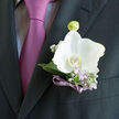 Pinned neatly above a suit pocket, this gentleman's boutonniere centres on a single white orchid that seems to glow against the charcoal fabric, like a calm light on a dusky Canonbury evening. The orchid's petals unfurl with gentle assurance, broad and velvety, touched at the outer edges with the faintest ivory warmth. Nestled around it are cool, silver-green leaves and delicate sprigs of lilac and gypsophila, their fine textures softening the composition and hinting at a garden in early bloom. A slender lavender-violet ribbon winds tightly around the stems, its subtle shimmer picking up the richer lilac tones of the tie beneath, so that flower, ribbon, and silk feel like a single, thoughtful gesture. The contrast between the pristine orchid and the dark, impeccably cut jacket is striking; it makes the flower appear almost weightless, as if it's floating just above the chest. You can imagine the wearer stepping out onto a quiet Canonbury terrace or making their way toward a ceremony near Upper Street, each step accompanied by the faint, fresh scent of newly cut flowers. The design feels measured and deliberate, as if chosen after careful conversation about style, colour, and meaning-something a seasoned neighbourhood florist would guide you through. This piece speaks of important days: a wedding morning full of soft anticipation, a formal celebration where family gathers, or a vow renewal marked by tenderness. In its refined simplicity, the boutonniere becomes a wearable keepsake of joy, grace, and the fleeting beauty of a perfect day.