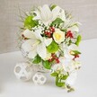 A refined, cascading white floral arrangement rests on a smooth white tabletop, offset by a soft beige, linen-like background that evokes a peaceful Canonbury dining room or boutique venue. The flowers are anchored in a round white woven basket, its tight wicker weave adding a handcrafted detail that matches the arrangement's natural elegance. Rising from this base, several large white lilies open generously, their long, trumpet-shaped petals gently curving back and revealing speckled throats and pale yellow stamens that suggest a delicate perfume. Cradled among the lilies is a single cream-ivory rose, fully formed yet still slightly cupped, offering a warm, romantic centre to the bouquet. From the right side, a graceful stream of white phalaenopsis orchids cascades downward, their broad, smooth petals and slender stems creating the impression of a floral waterfall, ideal for a wedding table near Canonbury's leafy streets. The arrangement is richly padded with greenery: wispy grasses, fine feathery foliage, and broader, glossy leaves that provide structure, depth, and movement. Cheerful bright green pom-pom blooms peek out like textured cushions, lending a modern, playful touch to the otherwise classic palette. Fine sprays of white baby's breath weave between the main flowers, creating clouds of softness, while small clusters of shiny red berries punctuate the design with bright, jewel-like colour, hinting at festive celebrations or winter weddings. To the left of the main basket, two decorative white spheres, crafted from interlaced strips into open lattice balls, sit quietly on the table, echoing the woven texture and adding sculptural interest. The whole arrangement feels luxurious yet calm, perfect as a wedding table centerpiece or special occasion display for gatherings across Canonbury.