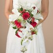 A close-up scene set in the heart of Canonbury captures a bride holding an immaculate white bouquet, with only her upper body and gently curved arms visible. This classic cascading arrangement, thoughtfully designed by local florists, features dramatic white lilies and trailing phalaenopsis orchids, both resplendent in the soft daylight that echoes the ambiance along Canonbury's leafy lanes. Enveloping these signature blooms are deep red roses-each petal lush and velvety-interwoven with whimsical baby's breath and eye-catching clusters of hypericum berries, whose bright red hues conjure images of winter market stalls by Canonbury station. The bouquet's texture is elevated by fern-like wisps and sturdy dark green leaves, providing a fresh foundation and highlighting the bouquet's flowing silhouette. Spiky green details add a contemporary twist amid tradition, while airy white filler flowers soften the composition. The bride's white gown, partially visible with subtle lace or satin detailing, emphasizes timeless elegance. A softly blurred beige background keeps the vibrant bouquet center stage, inviting the senses to linger on each flower's shape and hue. This bespoke bouquet, perfect for Canonbury wedding celebrations and receptions, reflects not just floral artistry but also the gentle romance and sense of occasion found across North London's storied neighborhoods.