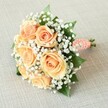 Resting upon a softly textured, neutral linen surface that echoes the understated elegance of Canonbury's townhouses, this lush bridal bouquet shines with expertly combined colors and thoughtful design. The centerpiece features around seven or eight large, peach-to-apricot roses at their highest bloom, their petals unfurling in graceful, spiraled layers that invite admiration. Clusters of delicate, cloud-like baby's breath nestle between each rose, orchestrating a dance of warmth and crisp white-an homage to dewy mornings beside the historic canal. Glossy, emerald-green leaves thread purposefully throughout, providing structure and a burst of garden vitality that grounds the bouquet's gentle romanticism. The compact, rounded silhouette is held together by a tactile coral-peach satin ribbon, its surface adorned with a meticulous spiral of tiny white faux pearls, glistening as if touched by the morning sun over Canonbury Square. The bouquet's stems, neatly trimmed and partially visible beneath the ribbon handle, hint at the freshness and quality expected from local florists. Even, soft lighting gently illuminates the entire composition, accentuating every curve and causing subtle shadows that add dimension and life. There's a palpable sense of poised joy-perfect for a wedding at St. Paul's Church or an anniversary celebration in one of Canonbury's hidden gardens. This bouquet, carefully curated for sophistication, speaks silently of enduring commitments and the unique community spirit that weaves through Canonbury's leafy streets.