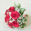 A classic, hand-crafted rose bouquet is laid out on a pale, textured linen-style cloth, its gentle beige tones offering a calm stage for the intense play of red, white, and green. In the centre, a close cluster of deep crimson roses, about six or seven blooms, opens fully yet gracefully, each velvety petal curling at the edges as if caught just at the peak of perfection. Between the roses, countless pinpricks of white baby's breath shimmer like tiny stars, the gypsophila forming frothy halos that soften the drama of the red. Surrounding everything, rich green foliage with smooth, slightly pointed leaves adds shape and structure, echoing the greenery along the quiet streets and leafy squares of Canonbury. The bouquet's stems are aligned in a neat bundle and wrapped in a vivid red ribbon, creating a comfortable handle that looks ready to be held by a bride stepping out from a townhouse near Canonbury Square or by someone delivering a romantic surprise. Along the length of the ribbon, small white bead-like details are arranged in a light spiral, adding a touch of sparkle and refinement without overpowering the flowers themselves. The overall arrangement is rounded and balanced, suggesting expert floristry and a sense of timeless ceremony. This image speaks of intimate weddings, anniversary dinners in local restaurants, or same-day rose delivery across Canonbury for meaningful declarations of love. The bouquet appears freshly tied, the roses plump and hydrated, inviting the viewer to imagine their soft fragrance and the gentle rustle of leaves as it is lifted from the fabric surface.
