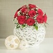 A compact, luxurious bouquet of deep crimson red roses sits gracefully in a modern white woven vase, evoking the refined calm of a Canonbury townhouse table laid for a wedding feast. Around a dozen to fifteen roses, each fully open and perfectly shaped, are gathered into a tidy sphere, their velvety petals layered so closely that the blooms form a continuous blanket of colour. Between them, delicate sprays of white baby's breath emerge in soft clusters, like tiny stars scattered across a night sky, brightening the arrangement and adding a feather-light texture. A few slim hints of fresh green foliage peek through, drawing the eye deeper into the bouquet and suggesting the cool, leafy gardens tucked behind local terraces. The flowers are anchored in a round, bulbous white vase fashioned from interlaced strands that create an open, airy lattice, reminiscent of artisanal wickerwork. Its crisp, clean lines give the arrangement a contemporary edge while allowing the vibrant red to truly stand out. Resting on the white tabletop in front and to the left are two decorative balls crafted from pale woven strips, one larger, one smaller, echoing the vase's texture and bringing subtle visual balance. Behind, a gently textured wall in soft beige remains out of focus, bathing the scene in even, flattering light. Everything about the composition suggests intimacy and celebration-a centrepiece perfectly suited to a wedding reception, engagement dinner, or romantic Valentine's meal at home in N1, where thoughtful design and heartfelt sentiment come together.