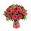A lavish bouquet of deep crimson red roses fills the frame, every bloom tightly clustered to create a luxurious dome of velvety petals. Each rose head is perfectly shaped, with petals that spiral from a tight, mysterious centre into wider, gently unfurling edges, giving a sense of fullness and depth. The colour is a true, saturated ruby red-rich, romantic, and dramatic-evoking passionate declarations and timeless love. Around and between the roses, glossy, dark green foliage, likely Ruscus or a similar leafy green, adds structure and contrast, its smooth, slightly pointed leaves catching the light and highlighting the roses' plush texture. The arrangement is gathered in a translucent ruby-red glass vase, whose twisted, spiralled surface design reflects and refracts light like ripples on water. The vase's flared rim and solid base give the bouquet a poised, elegant stance, while the rose stems, visible through the coloured glass, add depth and a natural, garden-fresh feel. Bright, even lighting falls over the entire composition, throwing every petal and leaf into crisp relief against a pristine white background, so nothing distracts from the drama of the flowers. The image feels like a romantic scene set for an anniversary dinner in a Canonbury townhouse, or a bold Valentine's gesture delivered to a flat near New River Walk-a bouquet that suggests heartfelt sentiment, celebration, and refined taste, without any extra decoration to dilute its message.