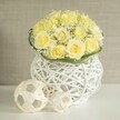 A serene, contemporary floral centerpiece is arranged with the care of an experienced Canonbury wedding florist, blending romance with clean design. Dominating the composition is a lush dome of soft, pale yellow roses, each bloom unfurled to reveal layers of velvety petals in tones of fresh cream and warm butter. They nestle close together so that the overall impression is one of fullness and luxury, without appearing heavy. Tucked among the roses, stems of white baby's breath introduce a fine, frothy texture; their minute blossoms scatter light like tiny stars, creating a gentle contrast that softens the structure. At the base of the bouquet, a neatly wrapped band of green foliage, perhaps woven leaves or slender blades of grass, encircles the stems, adding a crisp, botanical edge that quietly frames the flowers.   

Supporting this floral dome is a distinctive white woven vase, sculpted from smooth strands that crisscross and loop in an open, basket-like pattern. The vessel's shape subtly echoes a modern urn, broadening slightly at the top, narrowing at the centre, then widening again towards the base. Its airy lattice allows delicate shadows to play across the bright white tabletop below, reminiscent of dappled light under the trees near Canonbury Gardens. To one side, two decorative spheres crafted from pale, natural-looking strips of wood or rattan continue the theme of open weaving, their different sizes introducing a sense of gentle movement and balance. The larger sphere sits closer to the viewer, with the smaller tucked just behind, like art objects placed carefully for a wedding reception or intimate dinner party. The background is a softly blurred wash of warm beige or light grey, with a subtle texture that never competes with the main display. Overall, the image conveys quiet celebration and timeless elegance, suitable for sophisticated wedding tables, engagement parties, or refined home decor across Canonbury and neighbouring Islington.