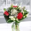 A contemporary mixed bouquet in a clear glass vase stands on a sunlit surface, captured as if in a bright Canonbury sitting room facing a large window. At the heart of the arrangement, a single coral-red rose opens generously, its velvety petals spiralling outward beside a ruffled crimson carnation, the two red blooms creating a strong focal point of passion and warmth. Around them, clusters of crisp white chrysanthemums and tiny daisy-like flowers soften the mood, their fine petals giving the arrangement a light, cloud-like texture. Creamy white lilies appear mostly in bud, their elongated forms adding height and a sense of anticipation, suggesting blossoms that will slowly open over the coming days. Deep emerald foliage is folded and arched around the base, the glossy leaves curving in wide bands that frame the flowers and give the bouquet a modern, architectural silhouette. The glass vase is simple and cylindrical, allowing the stems to be seen clearly, criss-crossing in the water and grounding the composition while reflecting the cool daylight. Gentle natural light, similar to the soft brightness along the quiet streets near Canonbury Station or New River Walk, washes over the bouquet, picking up every nuance of red, white, and green. The impression is one of fresh air and quiet celebration, with an imagined scent that combines the clean sweetness of lilies with the green, leafy notes of just-cut stems. This is the kind of arrangement that would sit beautifully on a dining table or sideboard in a Canonbury home, suitable for birthdays, thank-you gestures, or simply brightening an ordinary weekday.