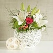 A sophisticated floral arrangement is captured in soft, even lighting, evoking the calm of a beautifully laid wedding table in Canonbury. The composition centres on a round, contemporary white vase, crafted from interlaced strips that form an open-weave, nest-like pattern, creating shadows and highlights along its curved surface. From this artistic container flows a generous bouquet of fresh blooms in white, red, and green, carefully balanced to feel both lush and light. Several prominent white lilies dominate the eye line; some are fully open, their graceful petals arching outward with a slight sheen, while others stand as smooth green buds, hinting at future blossoms and ongoing life. Among the lilies, velvety red roses add depth and emotion, their petals forming tight spirals that speak of romance and devotion, ideal for wedding decor across Canonbury and surrounding Islington streets. Bright green pom-pom flowers, likely dianthus or small chrysanthemums, punctuate the arrangement, their rounded forms and fine, bristling texture creating visual rhythm and a refreshing contrast. Tiny white flowers of baby's breath scatter delicately between the larger blooms, like a sprinkling of light, softening the transitions and adding an almost ethereal haze. Small clusters of red berries tuck into the foliage, introducing subtle seasonal warmth. The greenery below and around the flowers is abundant: slim, feathery fronds and broader, deeper green leaves intertwine to provide structure, movement, and a rich backdrop. The vase is placed on a pristine white surface that reflects hints of the arrangement above, and to the left two decorative woven spheres in a pale, natural wood tone echo the vase's textural theme. The neutral, lightly textured beige background fades quietly away, allowing every flower, leaf, and woven detail to stand out. Overall, the image conveys a sense of refined celebration and thoughtful craftsmanship, perfectly suited to wedding tables, engagement parties, or intimate gatherings in Canonbury homes and venues.