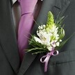 A close-up, high-angle photograph shows the upper torso of a man dressed for a formal event somewhere in Canonbury, his charcoal grey suit jacket filling the frame. The fabric looks smooth and finely tailored, with a clean, structured lapel line that catches the soft indoor light. Beneath the open lapels sits a pale dress shirt marked with a subtle grid pattern, its crisp cotton contrasting gently with the depth of the jacket. Running straight and true down the centre, a solid lavender necktie glows softly, the knot carefully shaped and sitting snugly at the collar. Pinned neatly to the left lapel is the true focus of the image: a handcrafted wedding boutonniere. At its top rises a slender, light green spire of tightly closed buds, perhaps hyacinth or muscari, suggesting fresh spring growth like you might see in nearby Canonbury Square Gardens. Around this are several open white alstroemeria blooms, each petal brushed with fine streaks of maroon or deep plum and revealing tiny golden stamens at the throat. Nestled between them, delicate sprigs of white gypsophila, or baby's breath, create a cloudlike softness, their tiny florets almost floating against the darker fabric. Slender, grass-like blades and slightly broader, glossy green leaves arc around the flowers, framing them with a natural, garden-fresh feel. At the base, a narrow satin ribbon in lilac-lavender, perfectly echoing the tie, is tied into a small, neat bow that gathers the stems together. The whole piece feels calm, refined, and quietly romantic, the kind of carefully considered buttonhole a groom or groomsman might wear for a Canonbury wedding, capturing both celebration and poise in one small cluster of flowers.