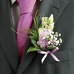 A close, carefully framed photograph shows the upper torso of a man dressed for a wedding or formal celebration, his charcoal-black suit jacket forming a dark, elegant backdrop for a refined buttonhole. The fabric of the suit has a soft, brushed texture, especially visible along the lapel edge and sleeve, while a neatly pressed, pale dress shirt with a faint grid pattern peeks through at the collar and cuff. At his throat, a bold magenta-fuchsia silk tie, smoothly knotted, catches the light with a gentle sheen, adding a playful flash of colour that feels right at home at a Canonbury registry office or a chic reception near New River Walk. Pinned carefully to the left lapel, a beautifully composed boutonnière becomes the true focal point of the image. Slender, still-closed buds of creamy white or pale snapdragon-like flowers rise gracefully at the top, suggesting blooms on the verge of opening. Below, a soft cluster of blush pink and lavender-lilac blossoms-reminiscent of sweet peas or delicate stock-nestles together, their petals slightly ruffled and velvety. Tiny white gypsophila flowers scatter like mist between the larger blooms, while a few richer purple florets add depth and contrast. Dark, glossy green leaves cradle the arrangement, giving it structure, while feathery tendrils of brighter green foliage arc outward, adding lightness and movement. Around the stems, a narrow lavender satin ribbon has been tied into a neat bow, its subtle gleam echoing the cooler purple tones in the flowers. The soft, natural daylight across the scene creates a feeling of calm joy and quiet anticipation, the kind of polished yet personal detail you might choose from a Canonbury florist to mark a wedding morning or special family gathering.
