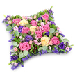 This image shows a luxurious, cushion-shaped floral tribute, designed for a funeral or memorial, resting serenely against a pure white backdrop. The pillow form is soft and rectangular with rounded corners, filled edge to edge with fresh flowers so that no part of the base is visible. A generous scattering of roses dominates the composition, ranging from vibrant fuchsia and deep pink to pale blush and gentle cream, their petals unfolding in delicate spirals. These are harmoniously paired with velvety lisianthus blooms in a rich royal purple, which thread through the arrangement and give it a softly regal air. Between the larger flowers, tiny lavender and lilac filler blossoms, much like statice or limonium, add intricacy and a sense of depth, ensuring every inch of the cushion is richly textured. Surrounding and supporting the blooms is a varied collection of greenery: silvery eucalyptus leaves, slightly rounded and matte, accompany darker, slender, fern-like foliage, together creating a cool green cradle that enhances the surrounding colour. The tribute appears freshly made, evocative of an early-morning delivery from a Canonbury florist preparing arrangements for a local chapel or crematorium service. Its calm pinks, creams, and purples suggest tenderness, sympathy, and peaceful farewell, much like the quiet atmosphere of Canonbury's residential lanes when families gather to say goodbye. Ideal for placing beside a coffin, at the head of a service, or on a graveside, this floral cushion visually communicates compassion and remembrance when it can be difficult to find the right words.