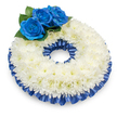 Seen from a gentle elevated angle against a clean white backdrop, this circular sympathy wreath is a study in quiet dignity. Hundreds of pristine white chrysanthemums are densely packed into a slightly domed band; their feathery, layered petals create a soft, cloud-like texture that catches a cool, diffuse light and throws a subtle shadow beneath the wreath, giving it gentle depth. Scattered centers of a warm, creamy yellow in a few blooms lend a trace of warmth to the overall cool palette. At the upper left, three deep royal-blue roses form a deliberate focal cluster - their velvety, tightly spiralled petals glow with a saturated sapphire tone and are tucked into dark green, rounded foliage that adds natural contrast and structure. Framing both the outer and inner edges of the chrysanthemum ring is a pleated satin ribbon in the same rich cobalt blue; the glossy ripples of the ribbon pick up highlights so the trim reads as both formal and tender, edging the piece with a refined sheen. The scent is understated: the soft, slightly honeyed perfume of the chrysanthemums barely lifting from the composition, offering calm rather than intensity. As a local Canonbury florist, I imagine this arrangement placed near a chapel in Canonbury Square or on a stand at a quiet memorial service along Essex Road - its palette and form are chosen for remembrance, purity, and solace. The design balances tradition with a personal note of blue, making it fitting for funeral services or as a consoling tribute to a loved one, conveying measured grief, respect, and enduring memory.