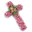 A softly coloured floral cross tribute in tones of blush pink, mauve, and gentle purple stands out clearly against a pure white background, with a faint drop shadow giving it a lifted, almost weightless presence. The entire cross is thickly carpeted with flowers, creating a luxurious, full texture that feels both comforting and refined. At the centre, three large roses in a dusty rose and light mauve shade form the heart of the design, their generous, layered petals opening with quiet grace. Around them, sprays of deep green foliage and feathery fern leaves weave between the blossoms, adding freshness and a hint of woodland softness. The arms and length of the cross are densely built from numerous lisianthus blooms, their gently ruffled, trumpet-shaped petals ranging from bright pink to richer, deeper tones, with some darker centres that add visual depth. Scattered throughout, tiny pink and purple accent flowers mingle with light green buds and the occasional small white blossom, introducing subtle variations in size and texture that keep the eye moving across the composition. Along the outer edges of the cross, slim glimpses of darker purple ribbon or fabric appear now and then, quietly framing the sea of petals. The design feels at once tender and dignified, the sort of tribute a Canonbury family might send to a local church or crematorium as a symbol of faith, remembrance, and affectionate farewell, offering beauty and solace when words feel hard to find.