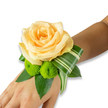 An intimate wrist corsage rests gently on the inner forearm, the skin a warm, light-to-medium tone that catches soft studio light, creating a calm, luminous backdrop for the flowers. At its heart sits a fully opened rose in a tender peach-apricot hue, its velvety petals layered in a slow spiral that invites a loving fingertip. Beneath the rose, two small lime-green button chrysanthemums-cheerful, almost spherical pom-poms-provide a lively, textural counterpoint; their tight florets and bright chlorophyll tone brighten the composition like a laugh beside a whisper. Deep, glossy leaves such as salal or ruscus form a dense, structured base, their rich green adding depth and a cool visual weight. A single variegated leaf, striped with soft cream and green and artfully folded into a sweeping arc, adds a modern, architectural flourish that guides the eye along the wrist and suggests motion when the wearer moves.

As a florist who often works in Canonbury and around Essex Road, I imagine this corsage worn at a local summer wedding at Canonbury Square or carried into a reception near Regent's Canal. The scent is a faint, elegant whisper of rose warmed by skin, underscored by a fresh grassy note from the chrysanthemums and foliage. The piece feels handcrafted, balanced between softness and structure-romantic yet contemporary-suitable for prom, an intimate ceremony, or a thoughtful honoured-guest gift. The fastening is discreet, likely an elastic or ribbon hidden beneath the leaves, leaving the floral artistry front and centre. Overall, this wristlet radiates warmth, celebration, and careful craftsmanship that complements both quiet mornings and evening festivities in Islington.