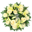 A quiet, dignified sympathy wreath captured from above, composed of a perfect circular base of deep green foliage cradling soft cream roses and pale yellow lilies. Laid flat against a clean white background and photographed in gentle, cool daylight, the arrangement reads as both refined and tender: roughly a dozen generously sized, fully opened ivory roses form the visual backbone, their tightly spiralled petals catching light like brushed silk. Between these blooms, trumpet-shaped lilies in a pale lemon tone sit at varying stages - some unfurled to reveal their soft stamens, others held in graceful buds - adding a subtle vertical rhythm and a whisper of hopeful renewal. The greenery is layered and textural: feathery fern fronds create a delicate halo at the wreath's outer edge while rounded, grey-green sprigs of eucalyptus or similar foliage nestle closer in, giving depth and a cool contrast that allows the creamy blooms to glow. The scent is imagined as a gentle, powdery sweetness from the roses with a faint, citrusy green of the lilies, a composition that feels both comforting and restrained. The wreath's circular silhouette, evenly balanced and densely packed, reads as a timeless symbol of remembrance and continuity - appropriate for a funeral service, memorial display, or graveside tribute. As a local florist who often arranges such tributes for families around Canonbury Square and nearby New River Walk, I picture this piece set with quiet respect in a chapel or beside a headstone, offering a measured, living consolation that speaks of purity, peace, and enduring care.