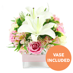 A modern yet romantic bouquet of white Oriental lilies and soft pink roses is displayed against a pristine white background, its colours bright and true as if lit by soft daylight streaming into a Canonbury flat. In the middle, the lilies open wide, their smooth white petals flaring outward to reveal dark reddish-brown stamens that add depth and contrast, giving the arrangement a graceful, sculptural centre. Around them, numerous pink roses cluster closely together, some petals still tightly coiled, others fully unfurled into generous, ruffled blooms, suggesting warmth and affection. Slender stems of pink and white alstroemeria, with inner petals subtly speckled in yellow and blush tones, weave between the larger flowers, while tiny clouds of white baby's breath lend a delicate, airy softness that lightens the whole design. The flowers are embraced by a rich frame of greenery, including shiny broad leaves and feathery fronds that add texture, movement, and the fresh feeling of nearby gardens along Canonbury Park. All of this is carefully arranged in a low, square clear glass vase, whose transparency reveals the neat stems and water, with a pale pink satin ribbon wrapped horizontally around its midsection to introduce a refined finishing touch. In the bottom right corner of the image sits a prominent circular orange label with bold white text that reads VASE INCLUDED, clearly indicating that this elegant container comes with the flowers. The scene conveys the impression of a thoughtfully composed, ready-to-display floral gift, ideal for birthdays, anniversaries, or simply sending comfort and beauty to homes and studios throughout Canonbury and the wider Islington area.